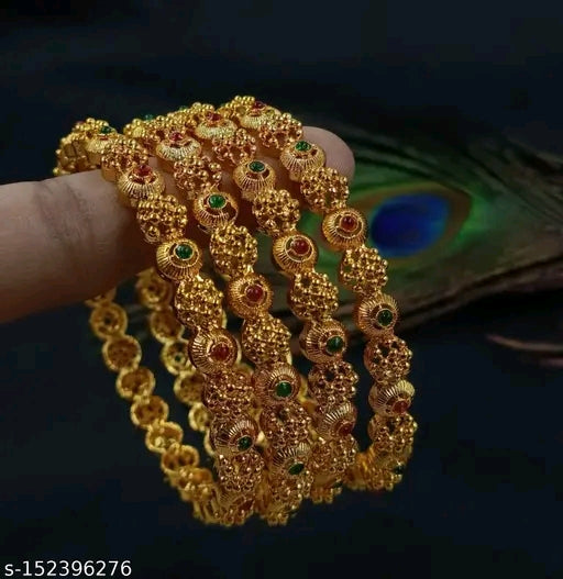 A series of gold-colored bracelets with artificial stones held in a person's hand, against a dark background with a peacock feather in the background.
