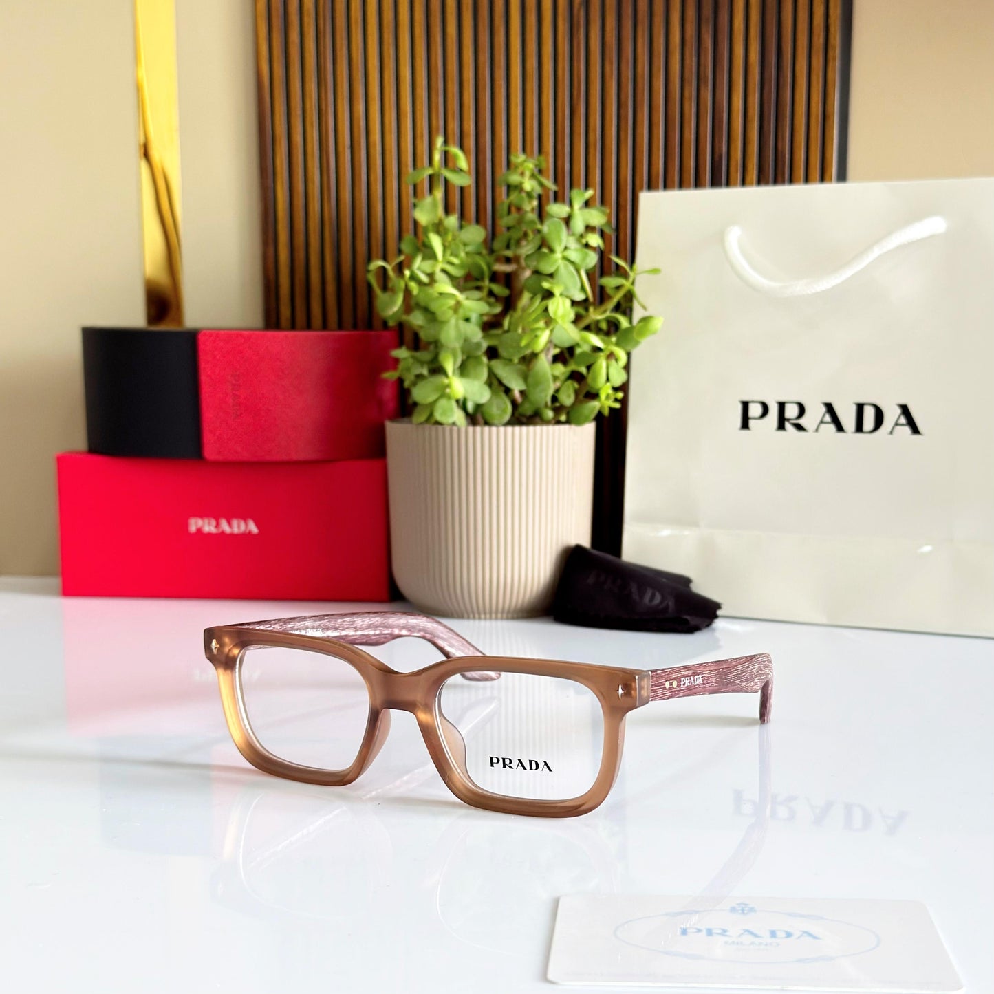 Brown Prada eyeglasses on a white surface with Prada packaging and a plant in the background.