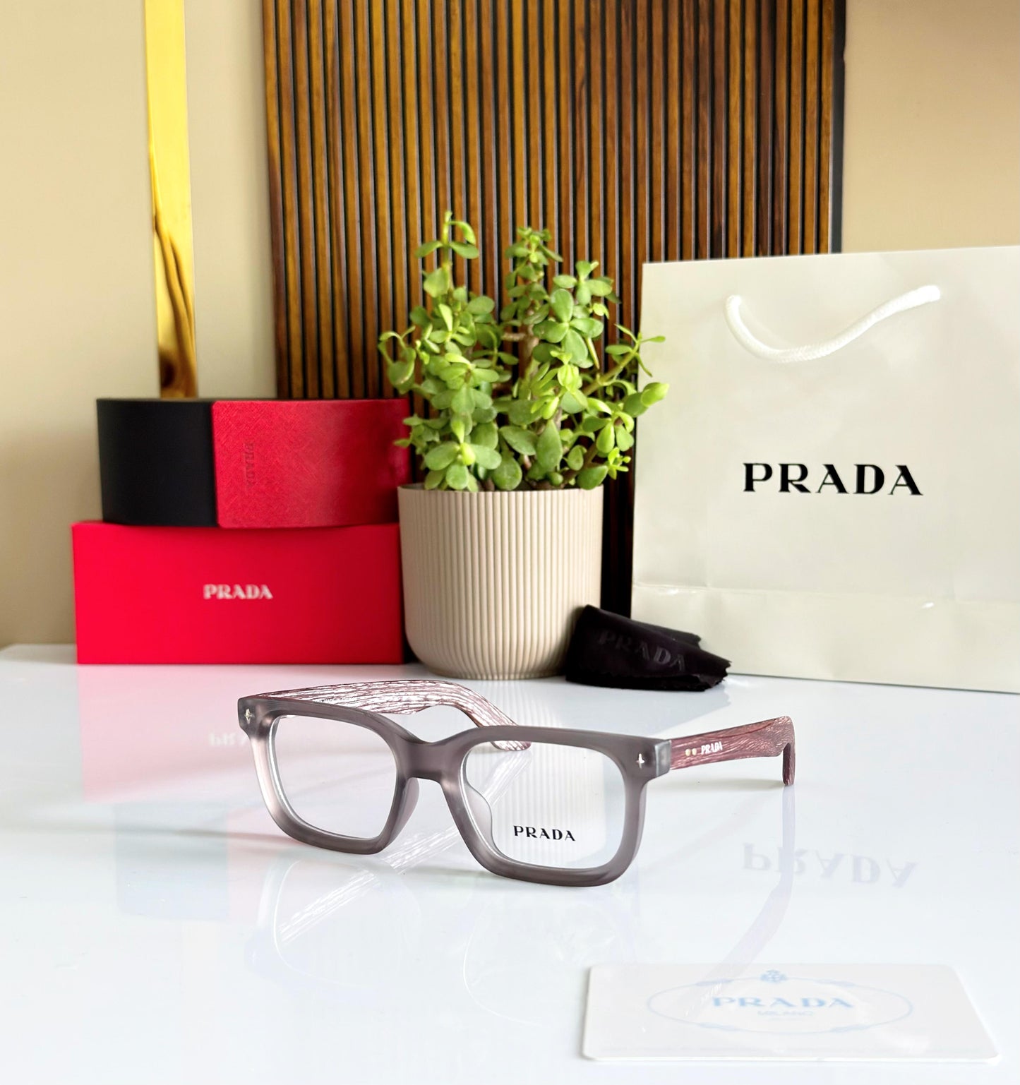Prada eyeglasses with red box, plant, and white Prada bag on a reflective surface.